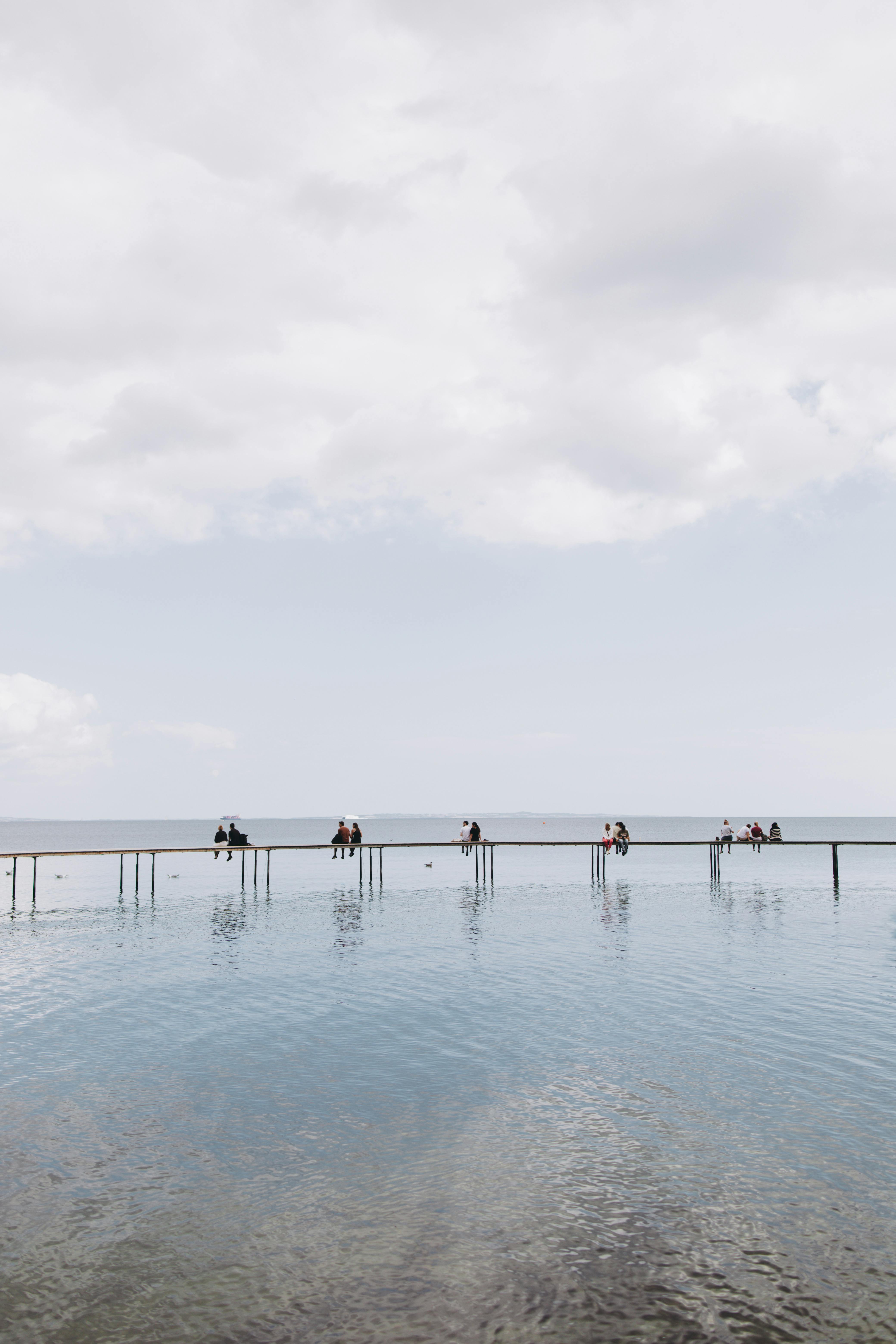 A serene scene of people relaxing on a long pier over calm water under a cloudy sky.