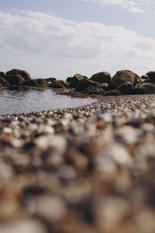Free A tranquil scene of a rocky beach with focus on pebbles and a blurred ocean horizon. Stock Photo