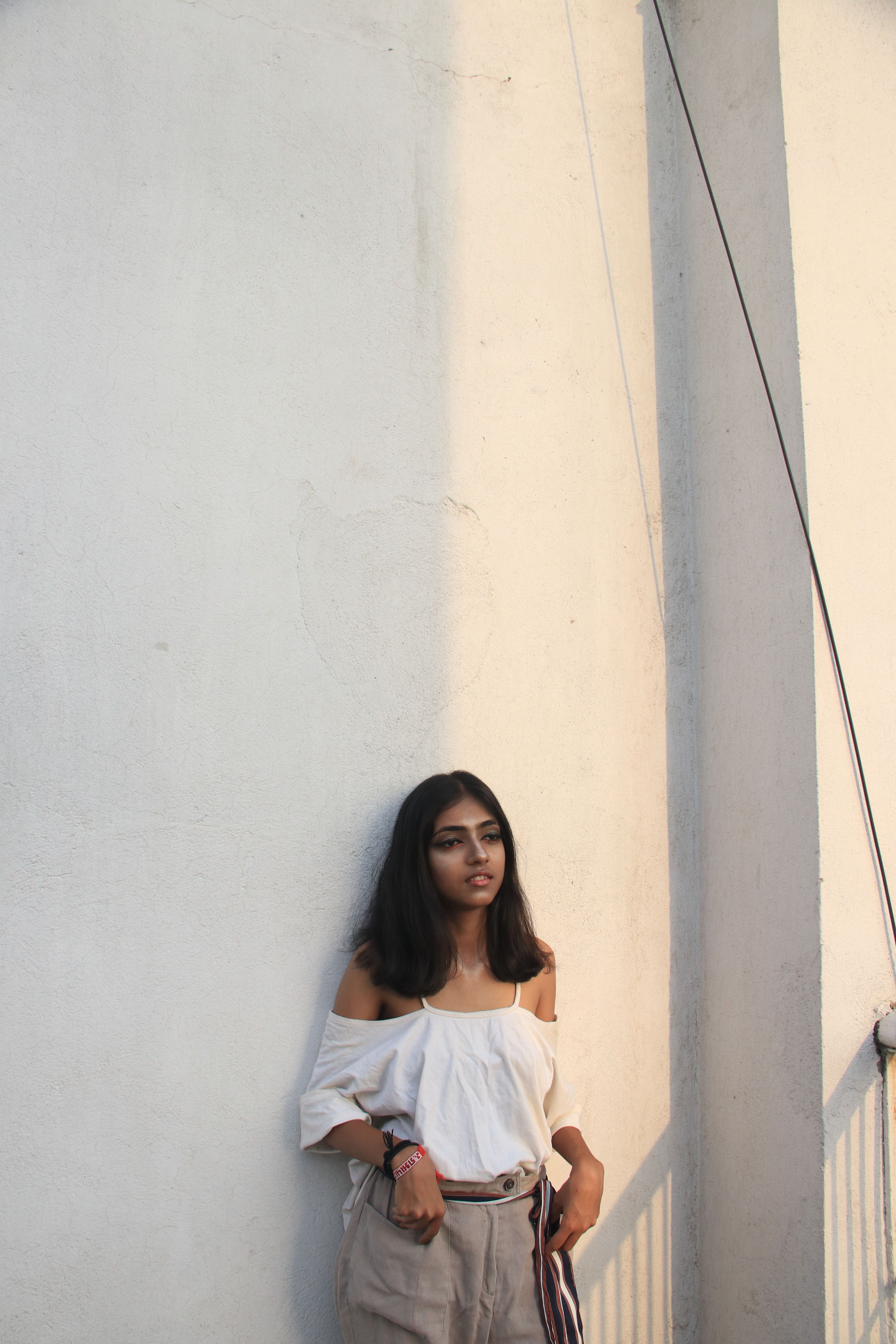 Woman in casual attire leaning against a sunlit wall, showcasing a relaxed and introspective pose.