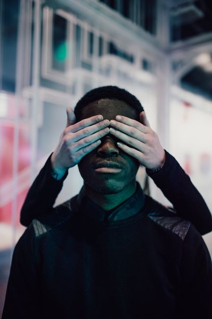 A man stands indoors with hands covering his eyes, creating a mysterious and playful scene.