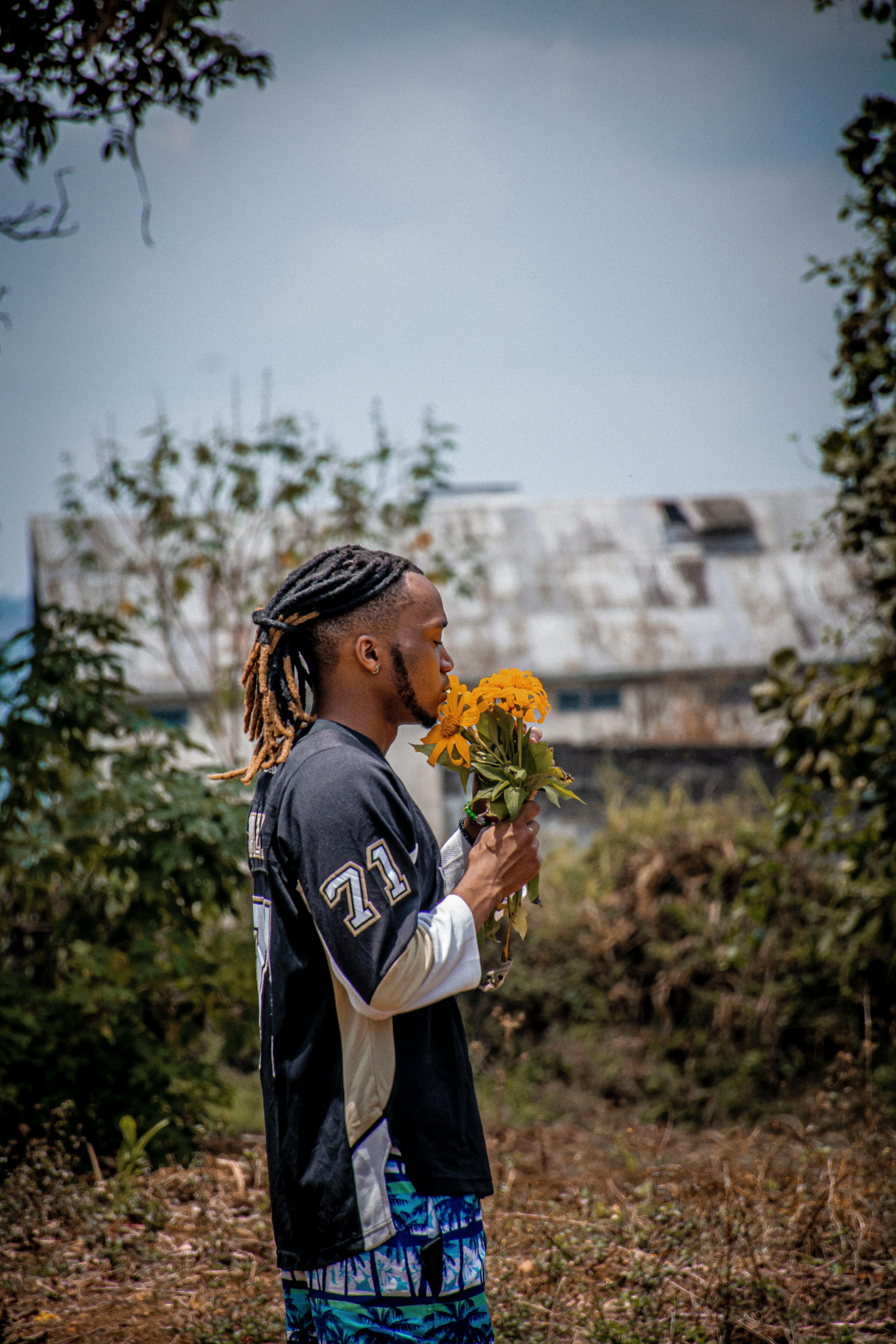 Free A man standing outdoors in a casual outfit holding a bouquet of sunflowers with a serene expression. Stock Photo