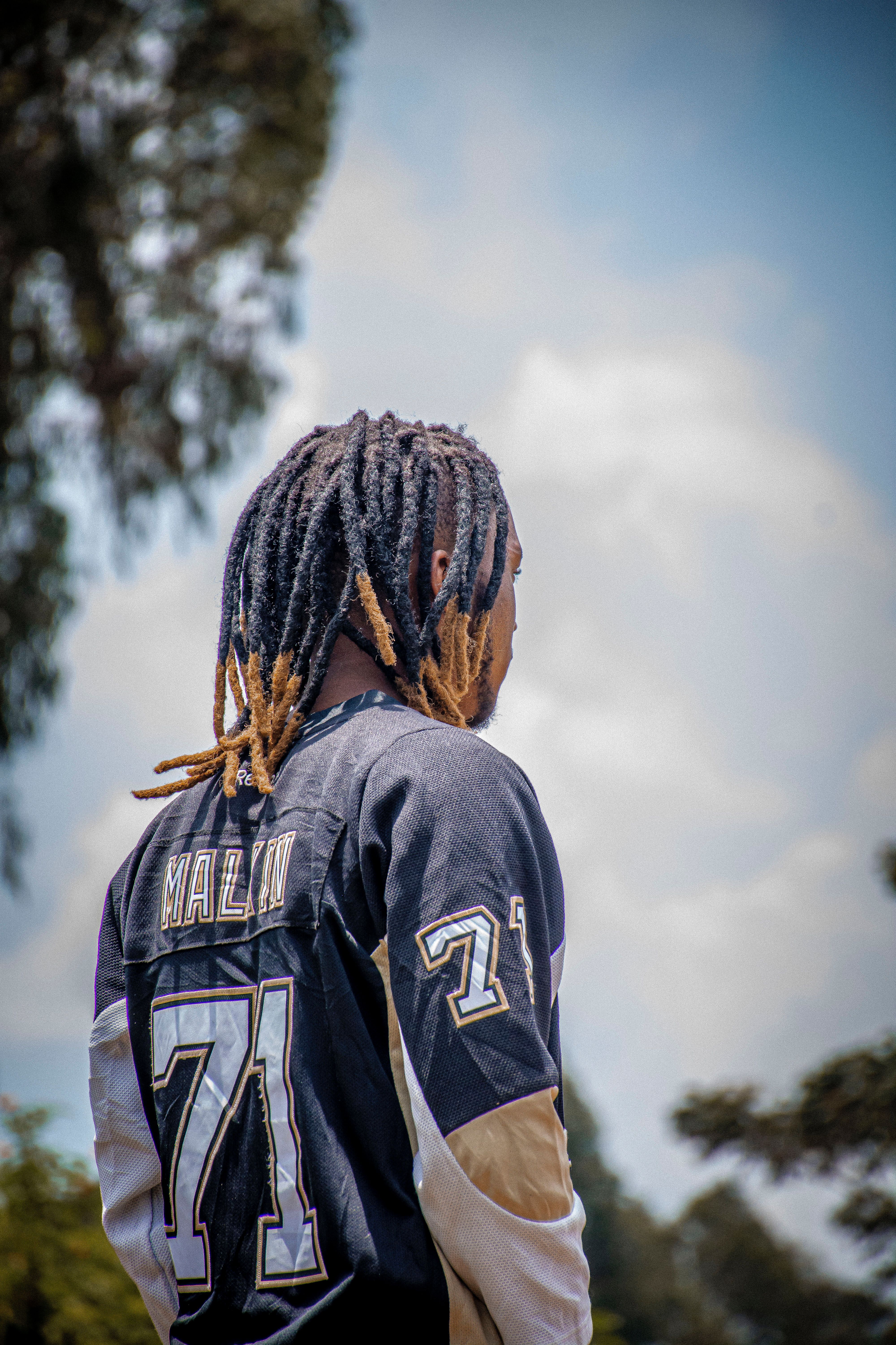 Man wearing sports jersey with dreadlocks, captured outdoors with a clear sky backdrop, conveying a sense of leisure and style.