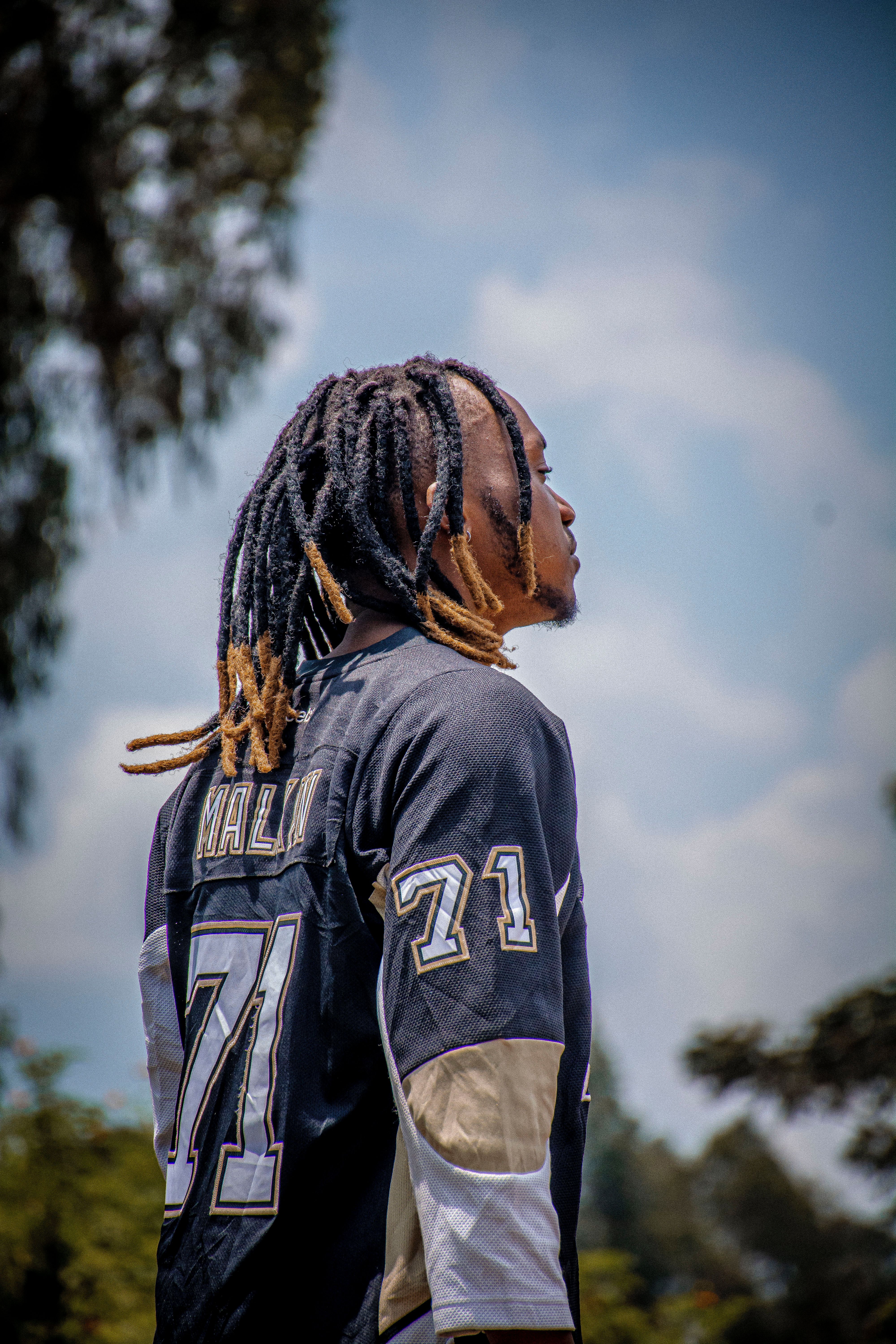 A man wearing a jersey looks upwards outdoors, showcasing his dreadlocks under a bright sky.