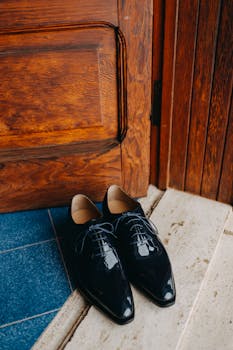 A pair of shiny black leather shoes placed near a wooden door on a tiled floor.