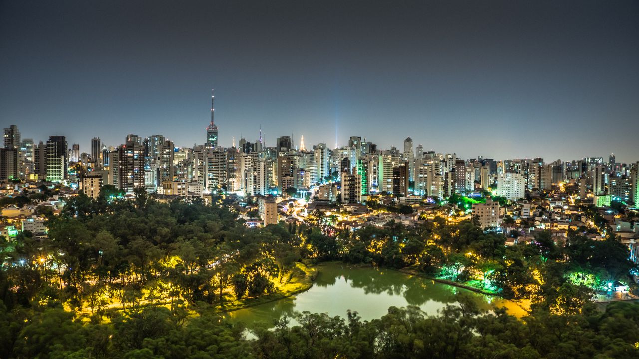 Night view of São Paulo's illuminated skyline with a park and lake in the foreground.