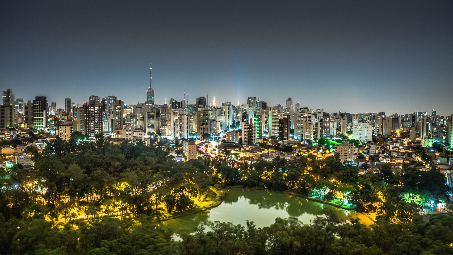 Night view of São Paulo's illuminated skyline with a park and lake in the foreground.