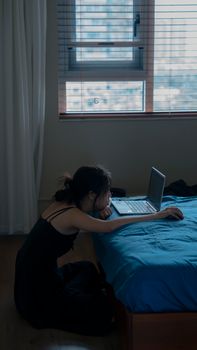 A woman sits on the floor of a bedroom, using a laptop on the bed under natural light.
