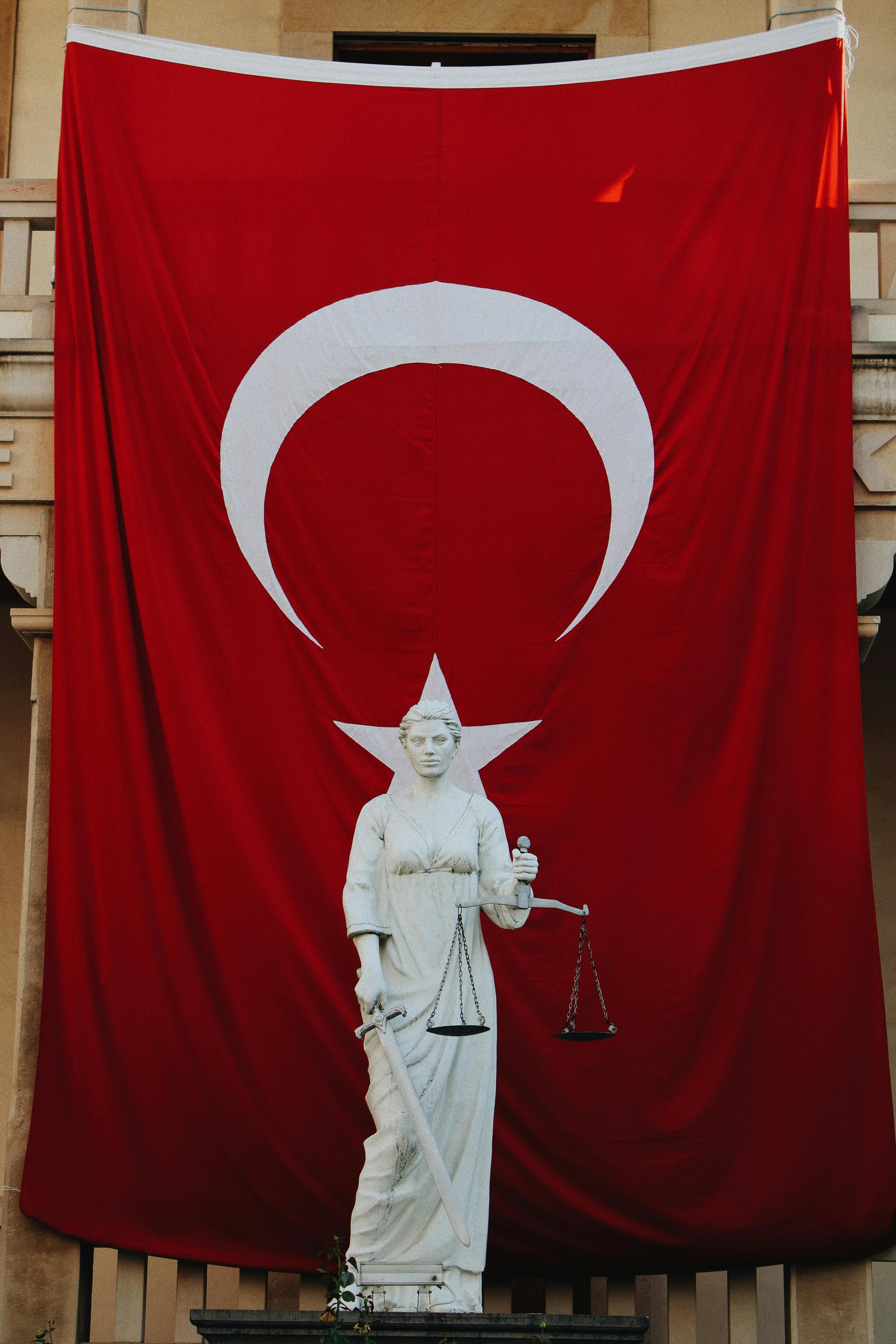 A statue of a woman holding scales in front of a large turkish flag