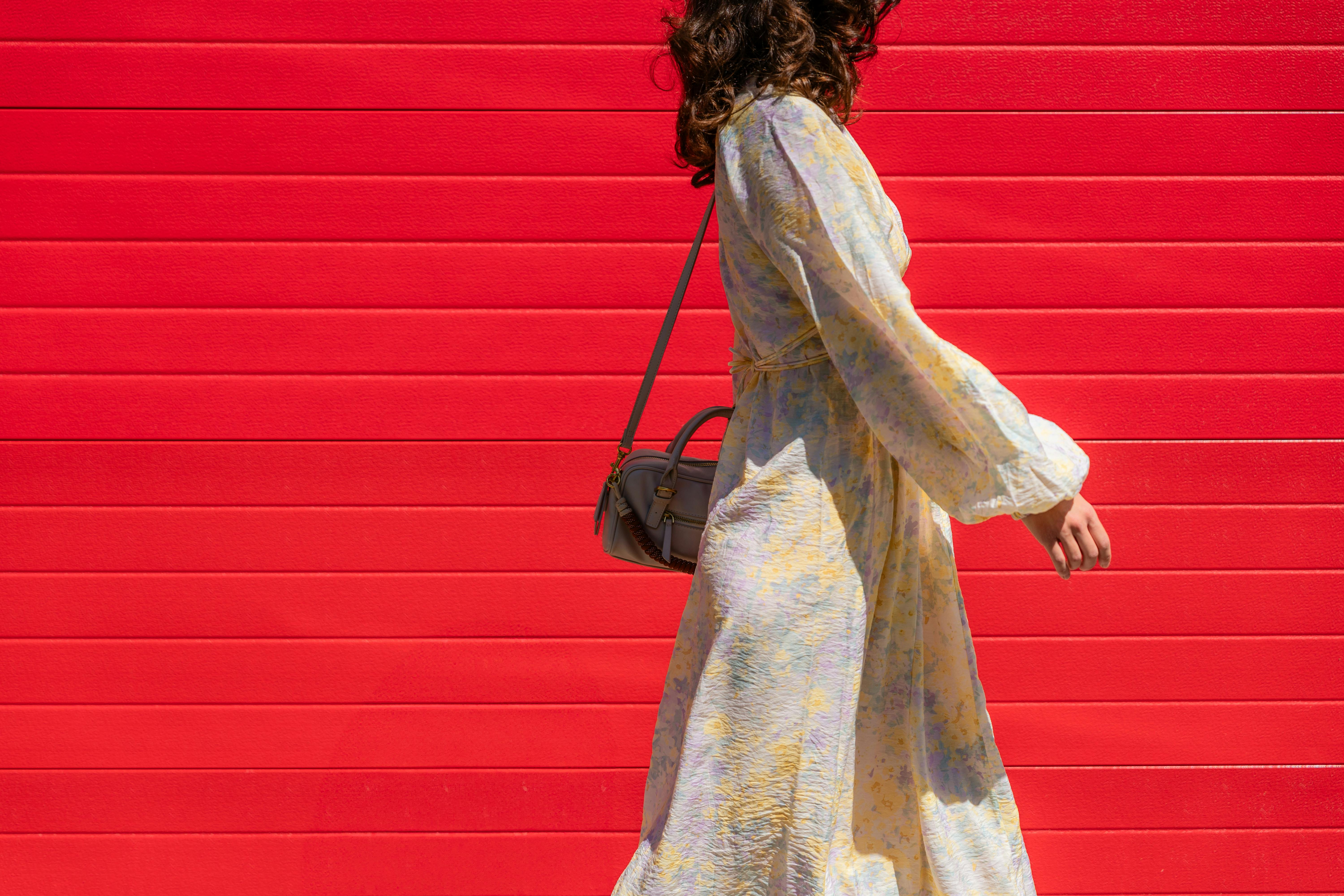Stylish woman walking past a bright red wall in a flowing dress. Captured in bright daylight.