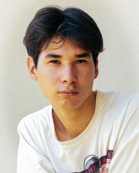 A close-up portrait of a young man with a serious expression, wearing a casual white t-shirt.
