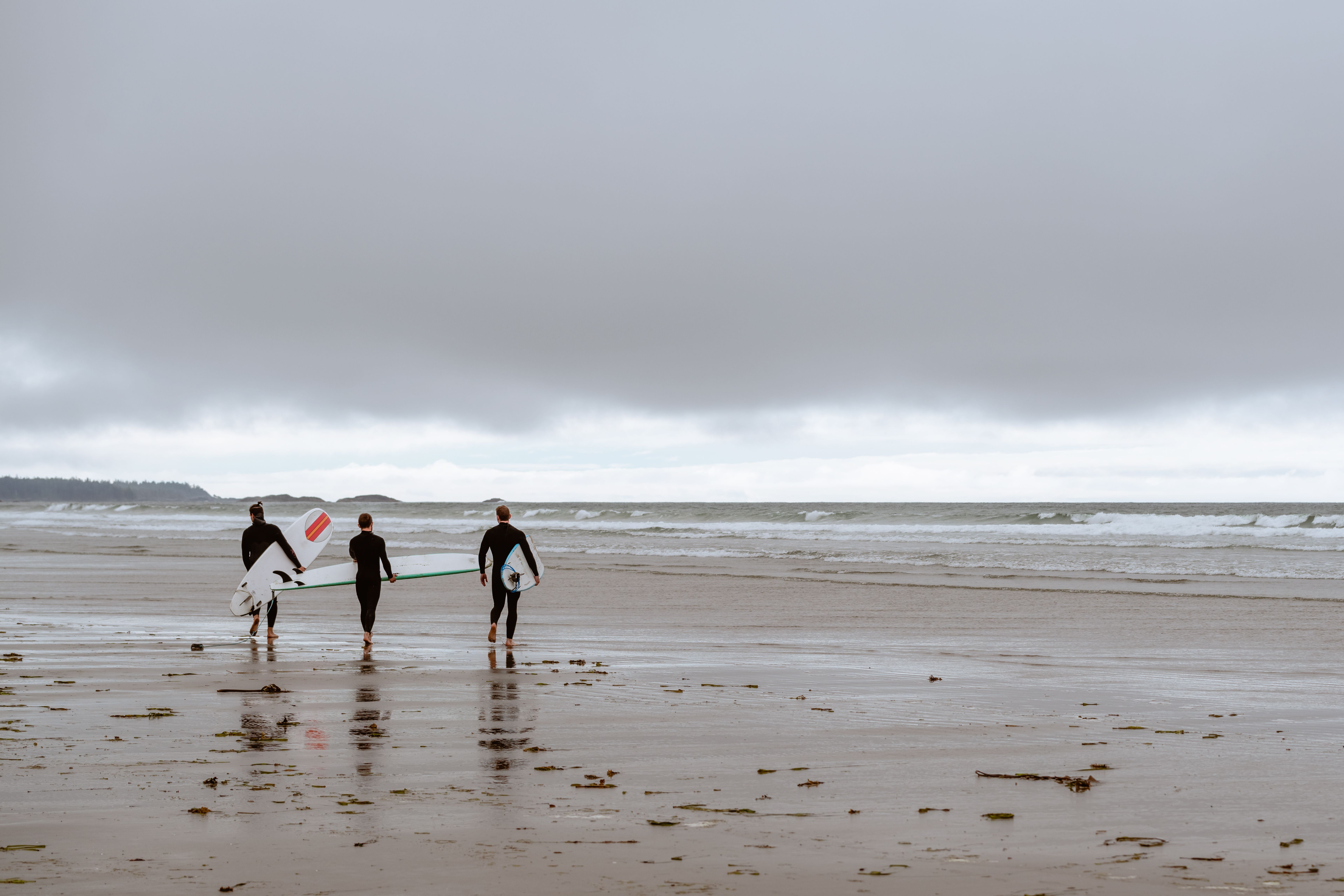 Three surfers walk along the beach in Tofino, BC, Canada, with surfboards on a cloudy day.