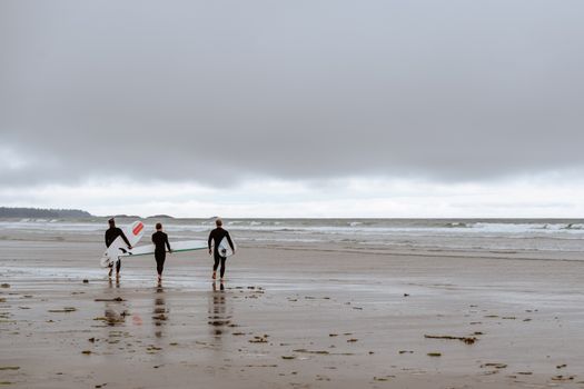 Three surfers walk along the beach in Tofino, BC, Canada, with surfboards on a cloudy day.