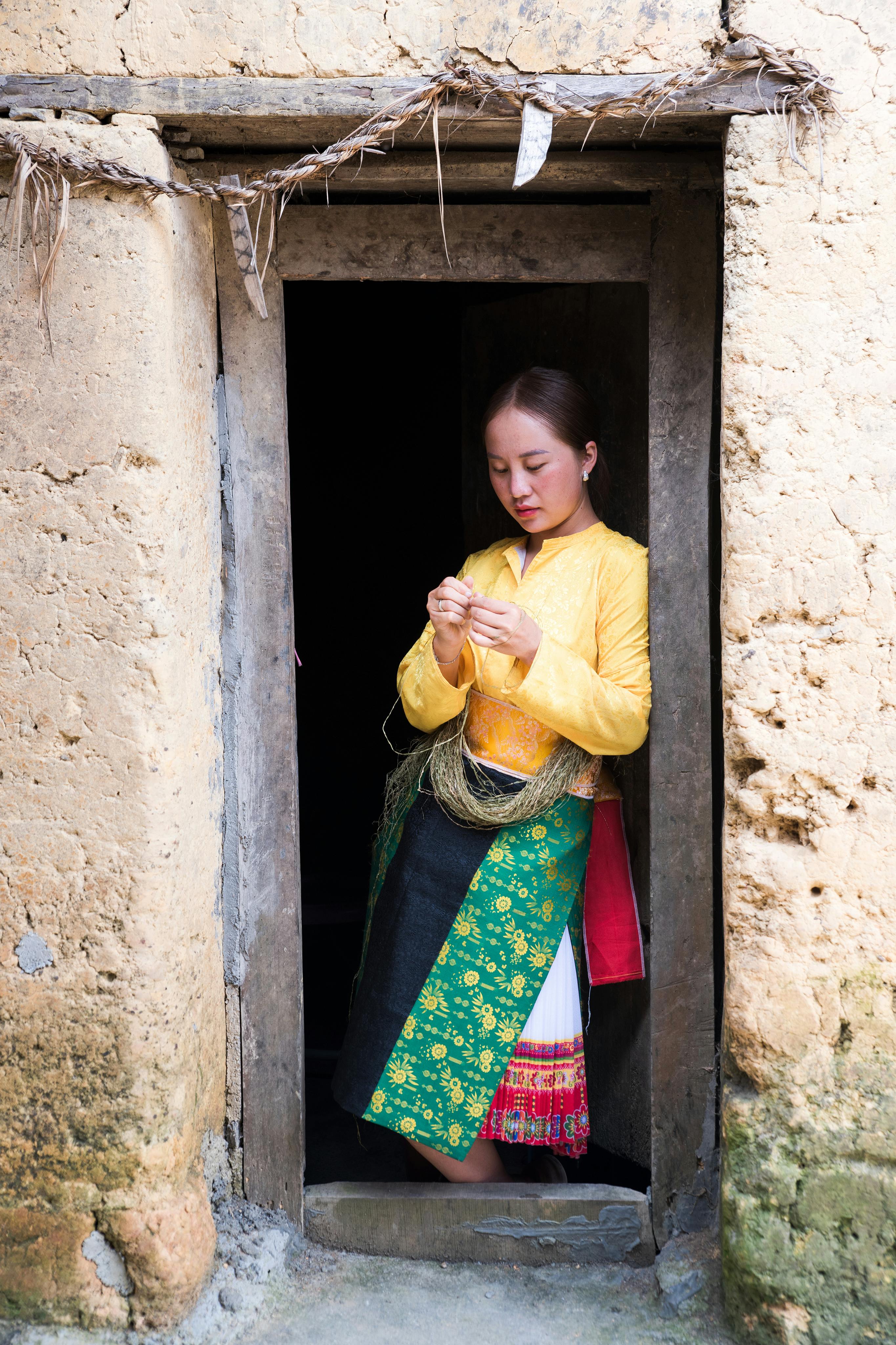 Woman in colorful traditional dress stands in rustic doorway, engaging in handcrafting.