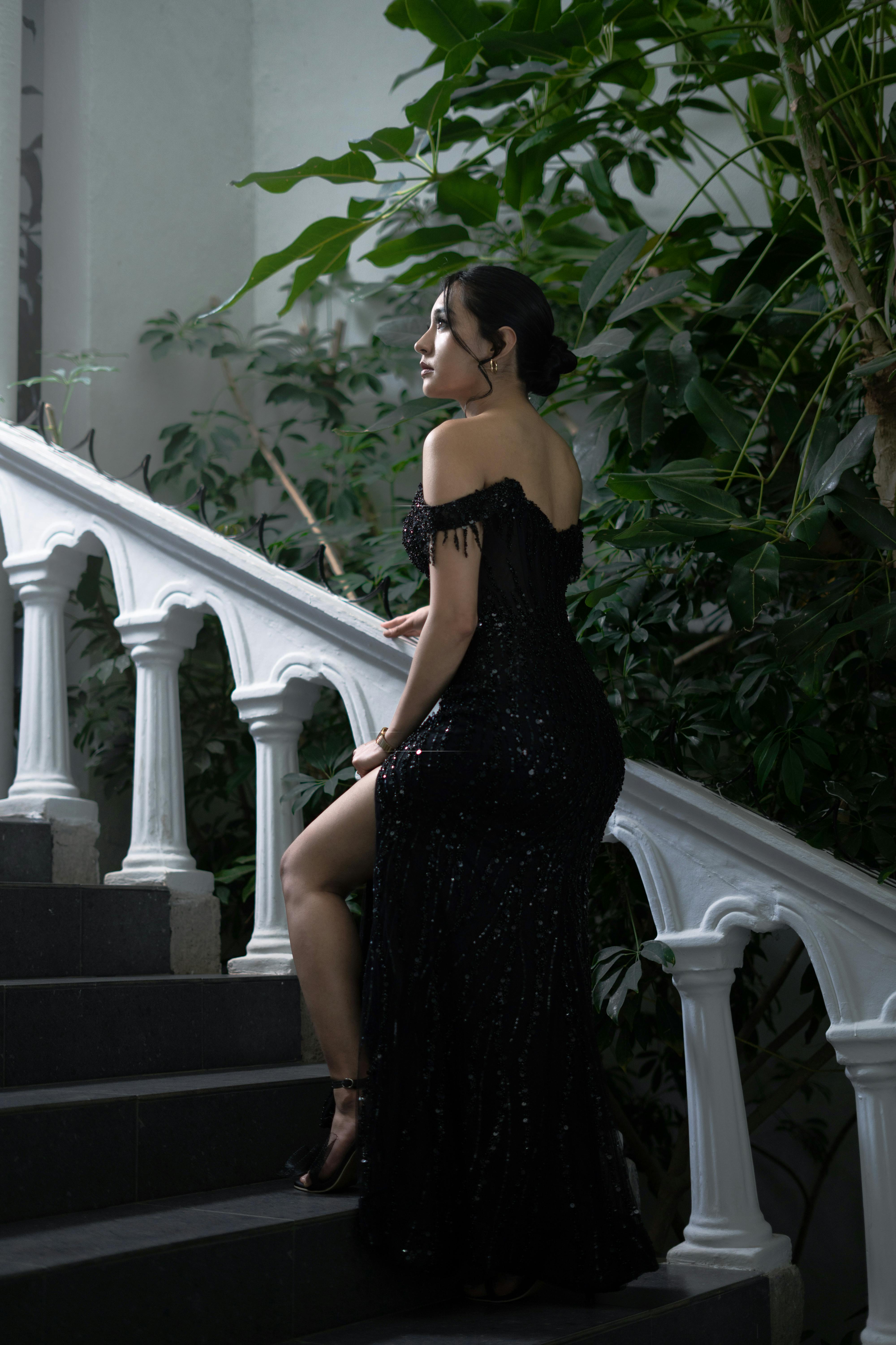 Elegant woman in a black gown on a white staircase surrounded by lush greenery.