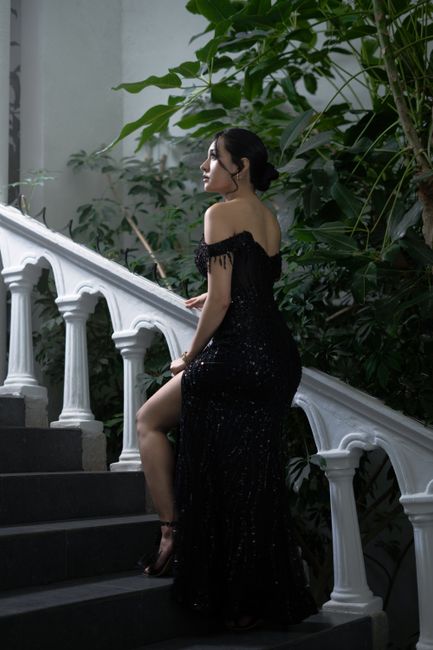 Elegant woman in a black gown on a white staircase surrounded by lush greenery.