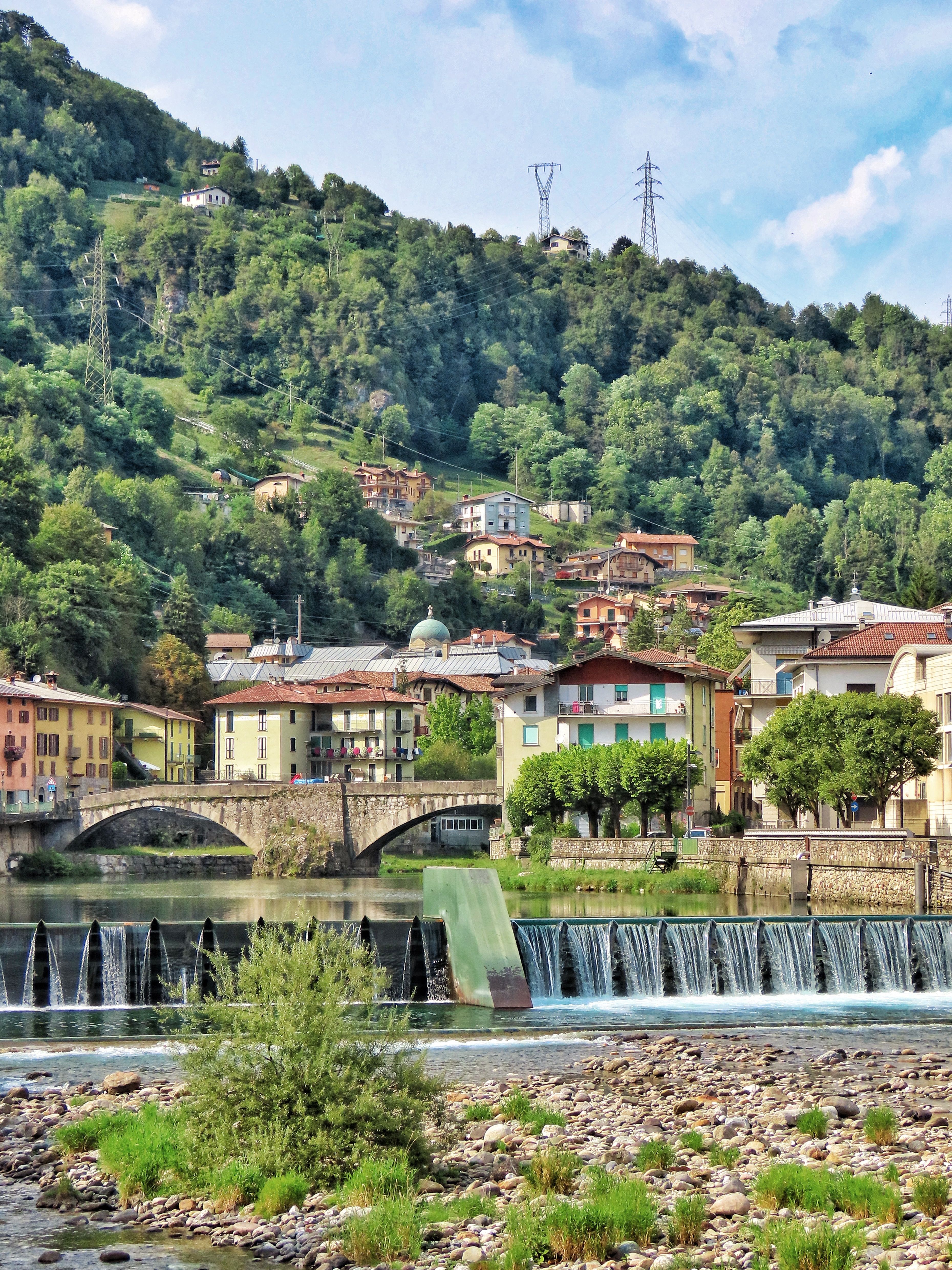 Charming riverside village with bridges, trees, and colorful houses in summer sunlight.
