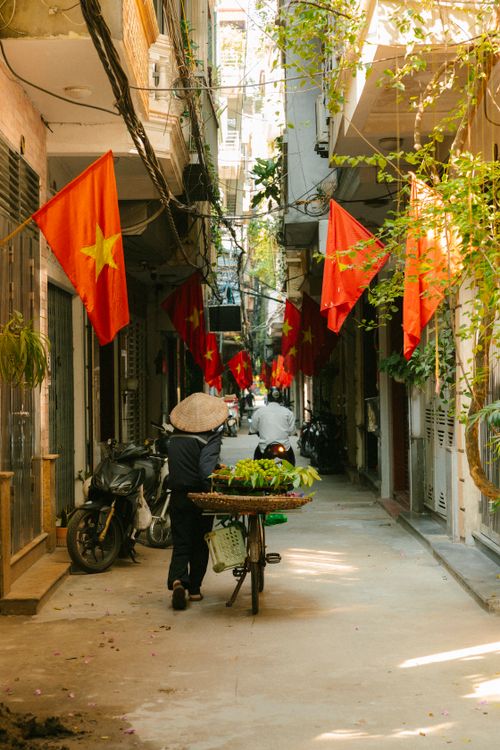 Free Sunlit alley with Vietnamese flags and vendor pushing a bicycle cart of greens. Vibrant everyday life in Vietnam. Stock Photo