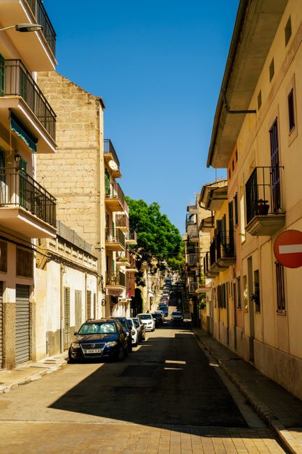 Picturesque narrow street in a Mediterranean town on a sunny day, lined with cars and balconies.
