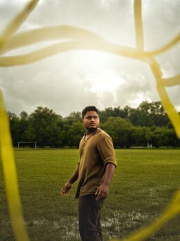 A man on a wet soccer pitch surrounded by green trees, under a cloudy sky.