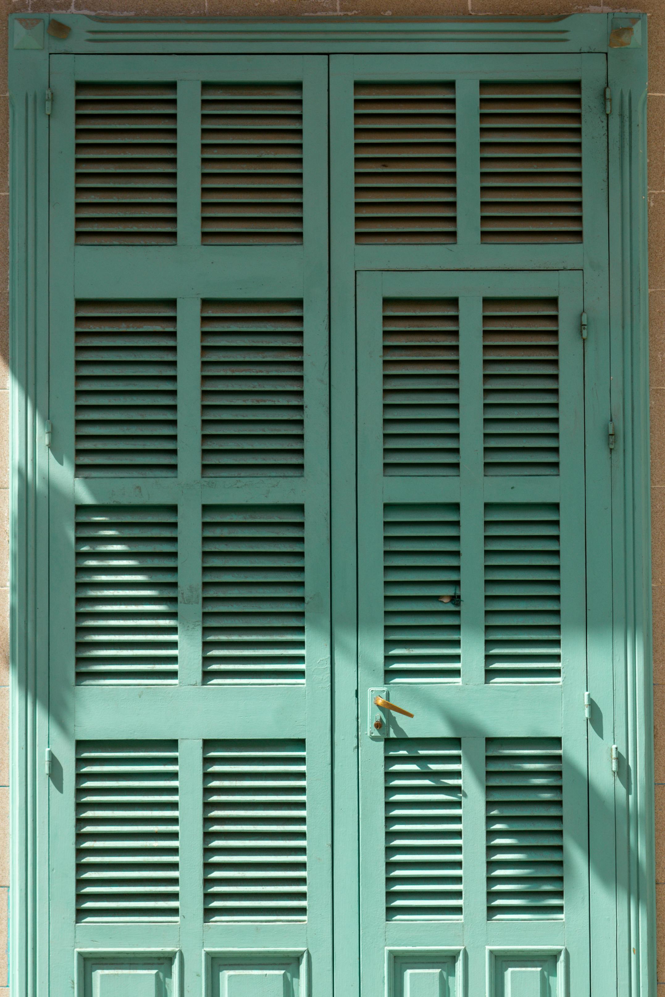 A vintage teal shuttered door with elegant wooden design and sunlight creating intriguing shadow patterns.
