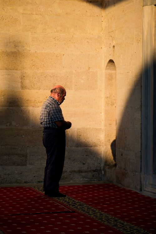 Free An elderly man stands in prayer, illuminated by sunlight inside a mosque, with shadows creating a dramatic effect. Stock Photo