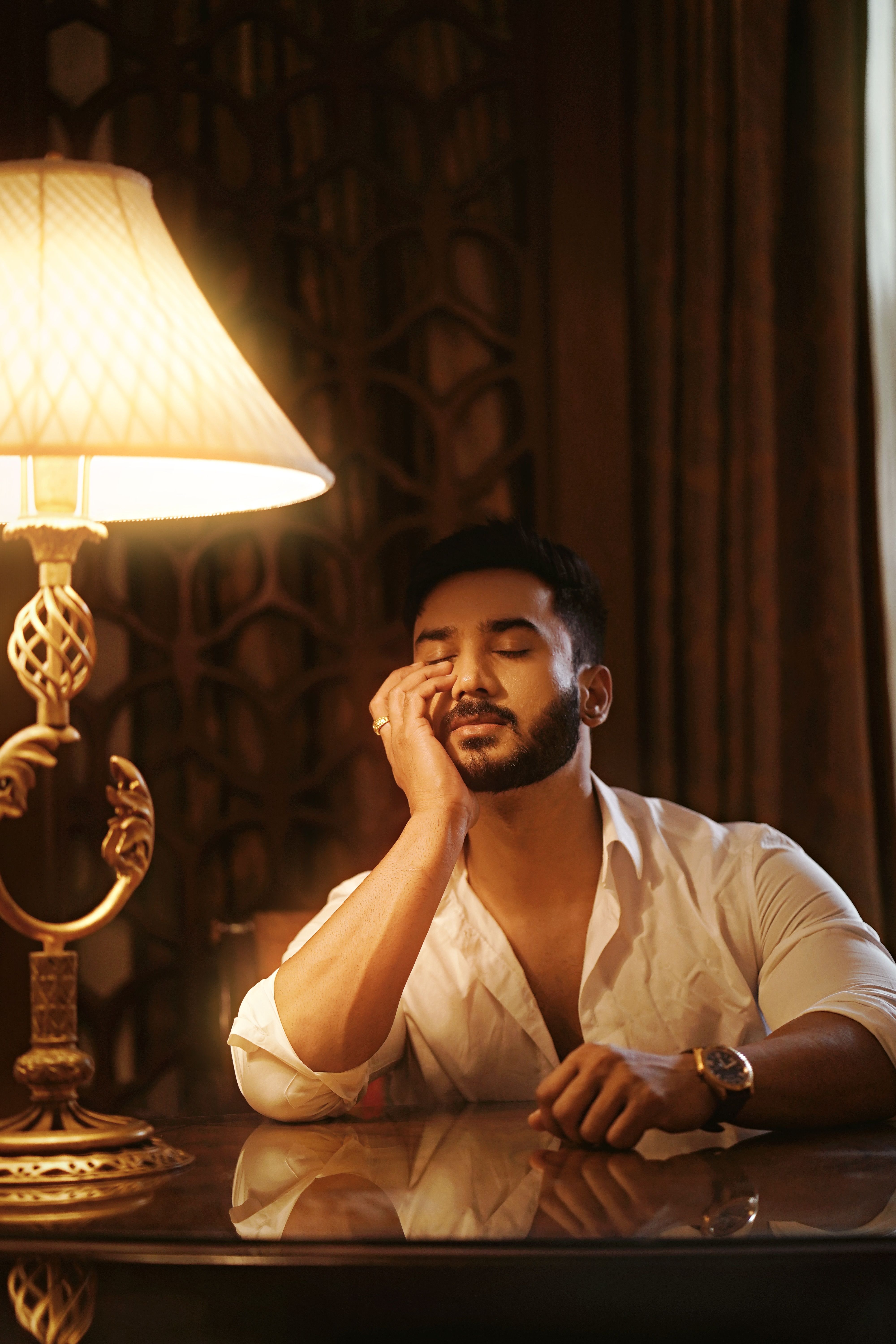 A man in a white shirt rests his head thoughtfully indoors by a warm lamp.