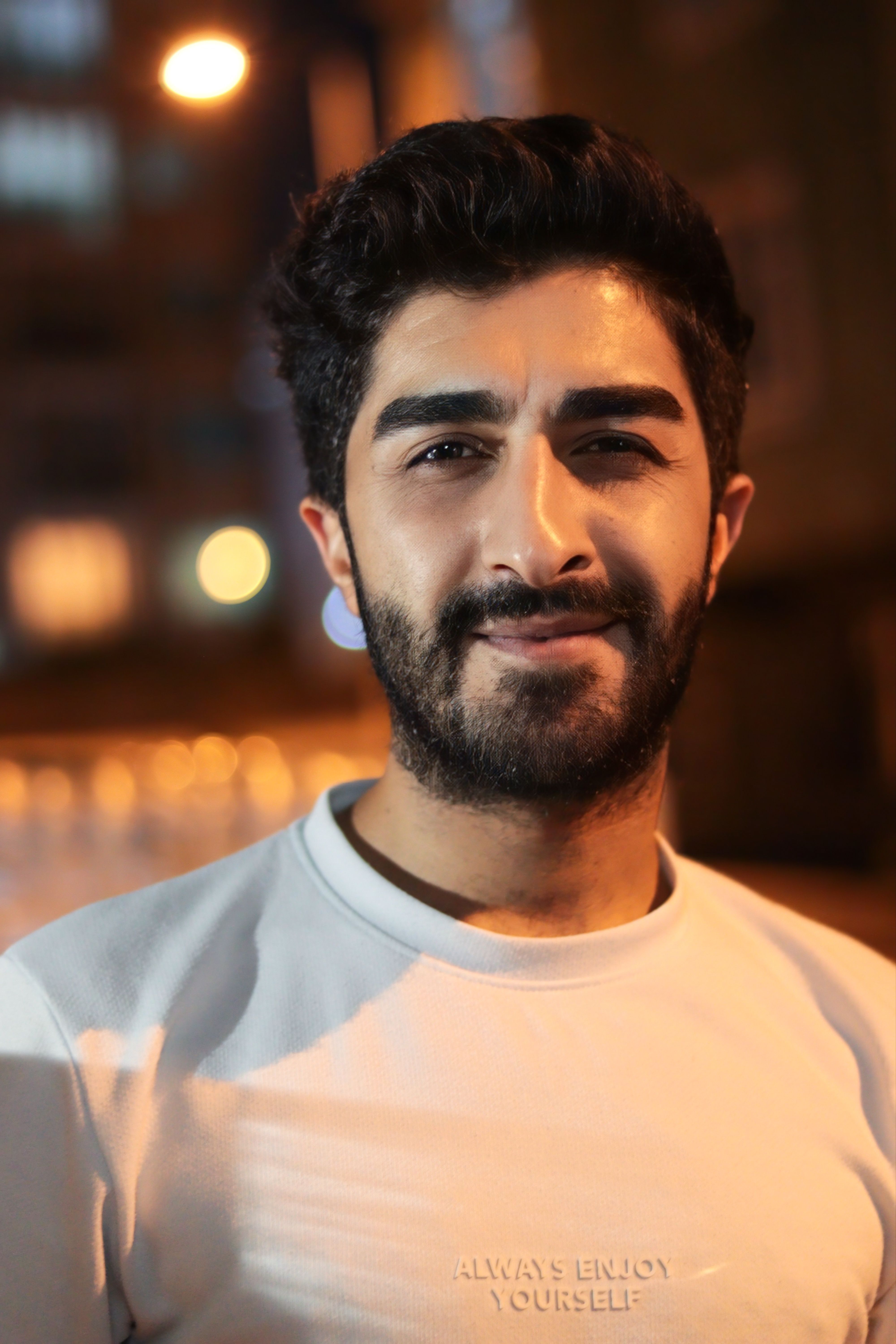 A striking portrait of a bearded man in casual wear, captured outdoors at night with warm lighting.