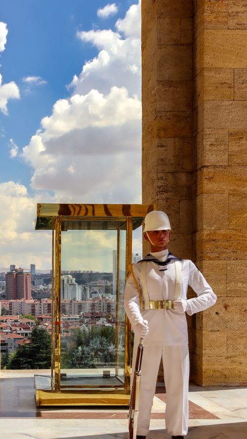 Free A soldier in white uniform stands guard at a monument with a cityscape background under a bright sky. Stock Photo