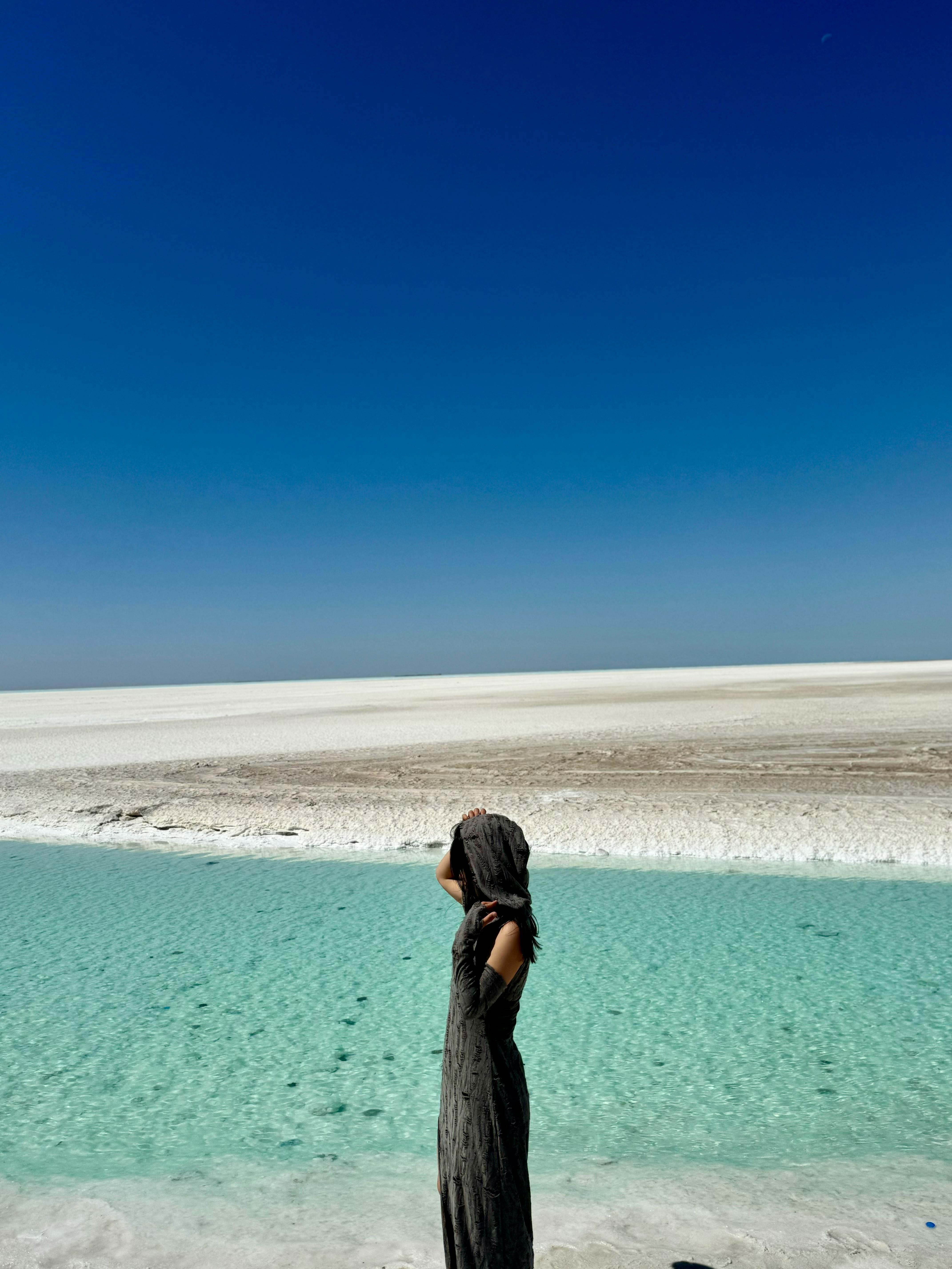 A woman in a headscarf stands by a serene turquoise lake under a clear sky, evoking tranquility.