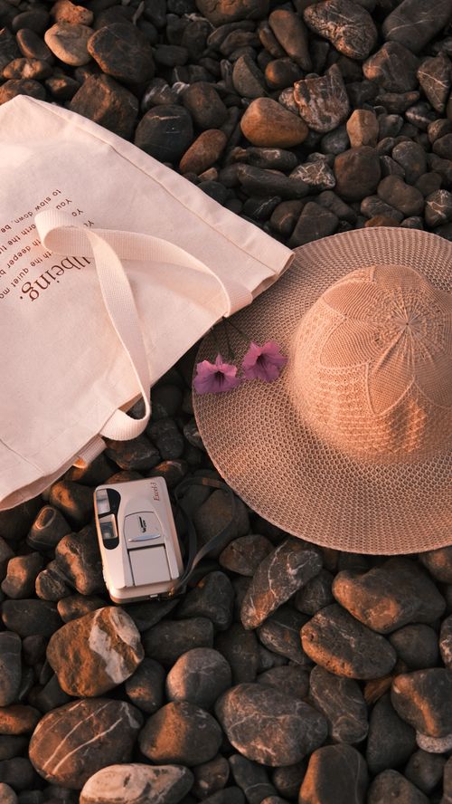 Free A peaceful flat lay of beach essentials including a hat, tote bag, and camera on pebbles. Stock Photo