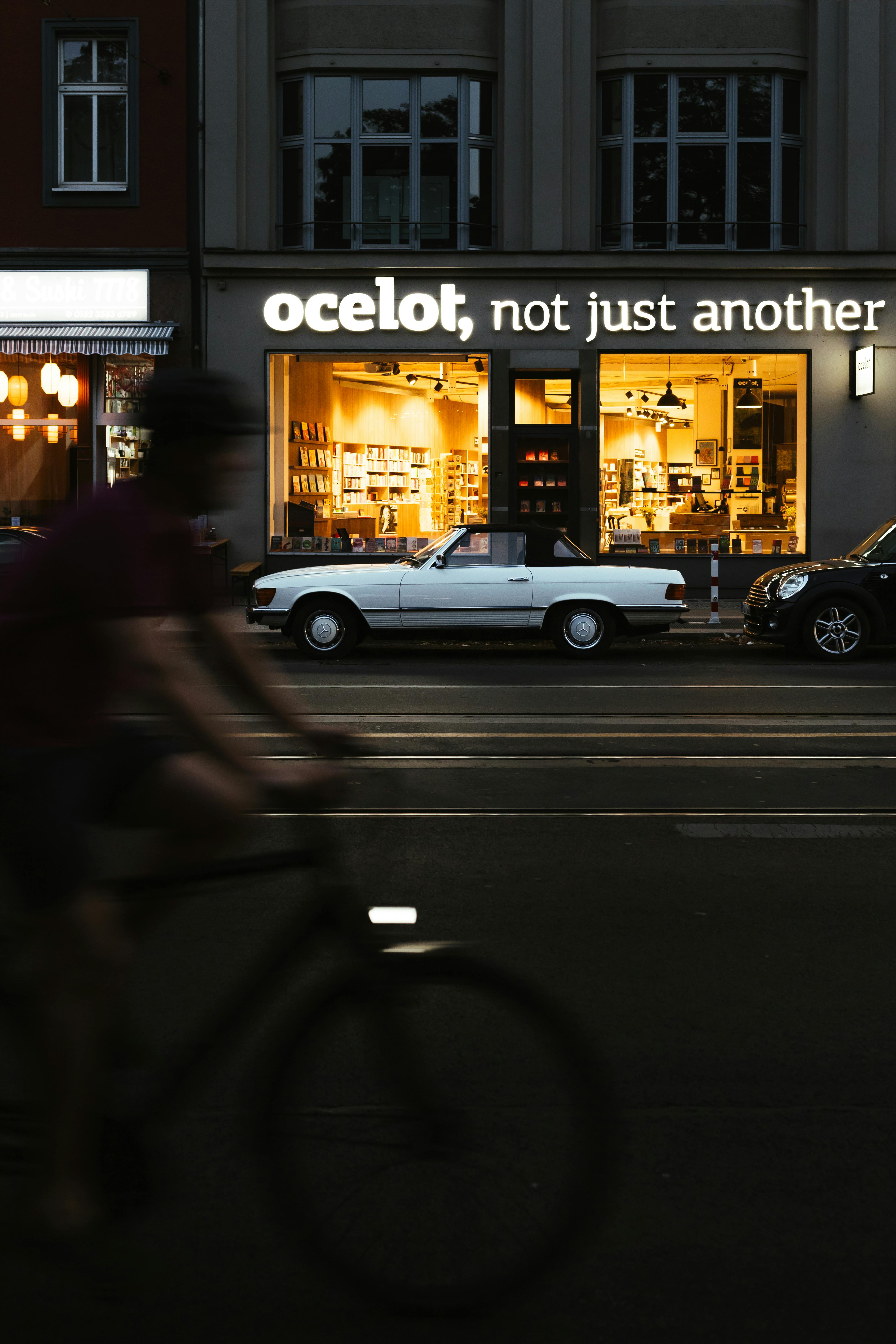 A moody evening street scene in Berlin featuring a cyclist and a car outside a well-lit store.