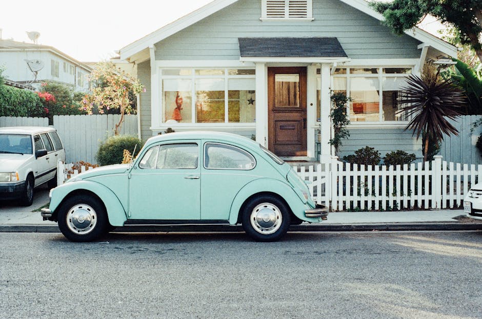This is a photo of a light mint green old VW beetle car in front of a wodden house with a white fence in a residential area in a typical american street. There is a yucca palm tree in the lawn of the house. The sun is shining brightly.