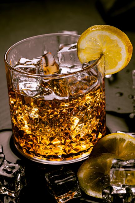 A Gold Rush cocktail in a rocks glass over a large ice cube, garnished with a lemon wheel, with a shaker and bourbon bottle in the background.