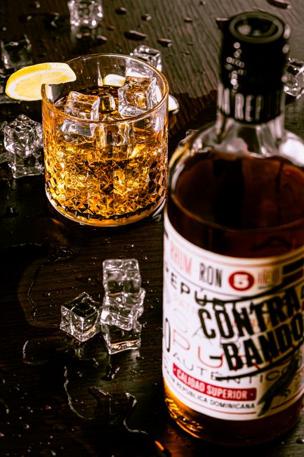 A bottle of clear white rum next to a glass with ice, lime wedges, and mint on a bar counter.