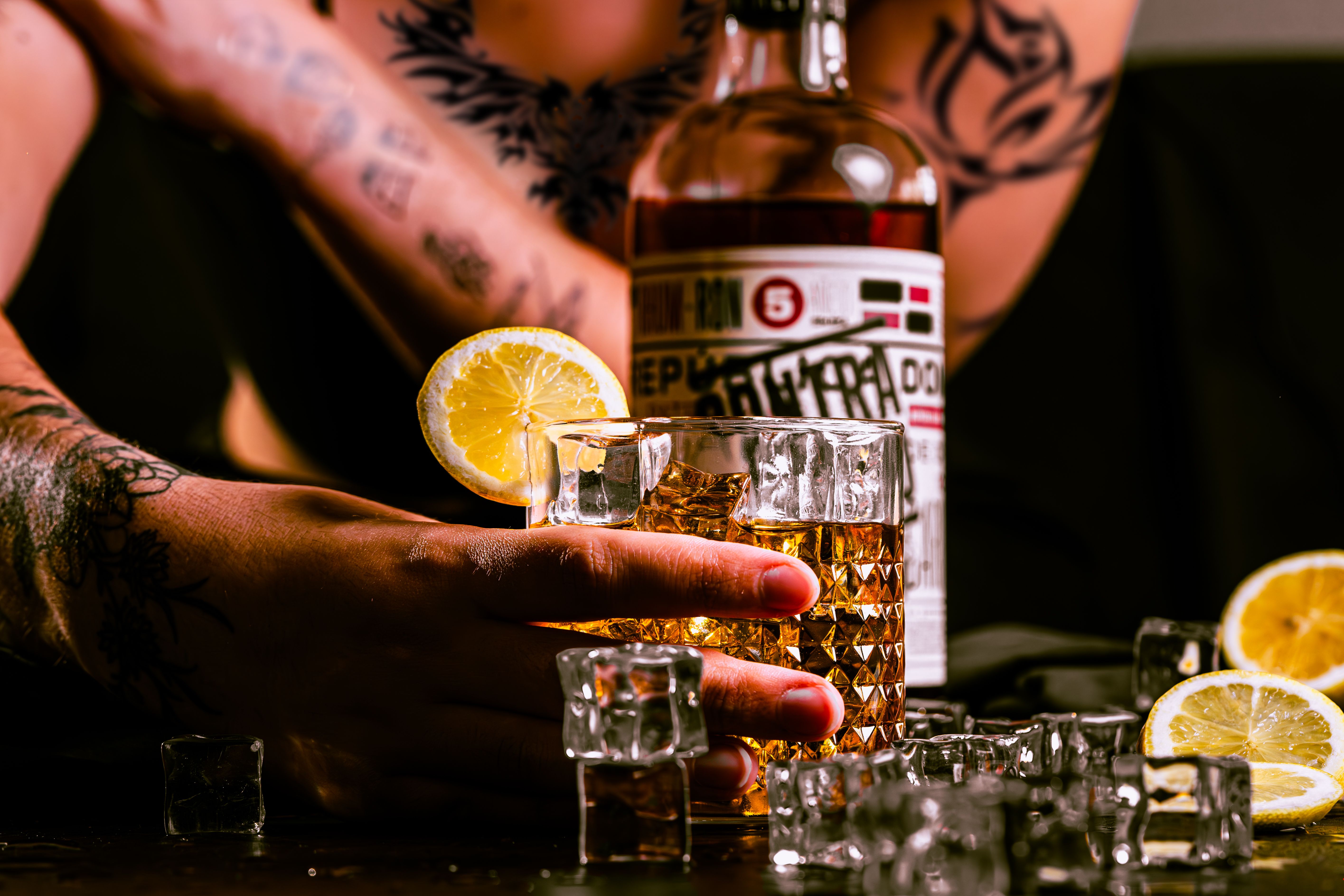 Close-up of a tattooed person holding a whiskey glass with lemon garnish in nightlife setting.