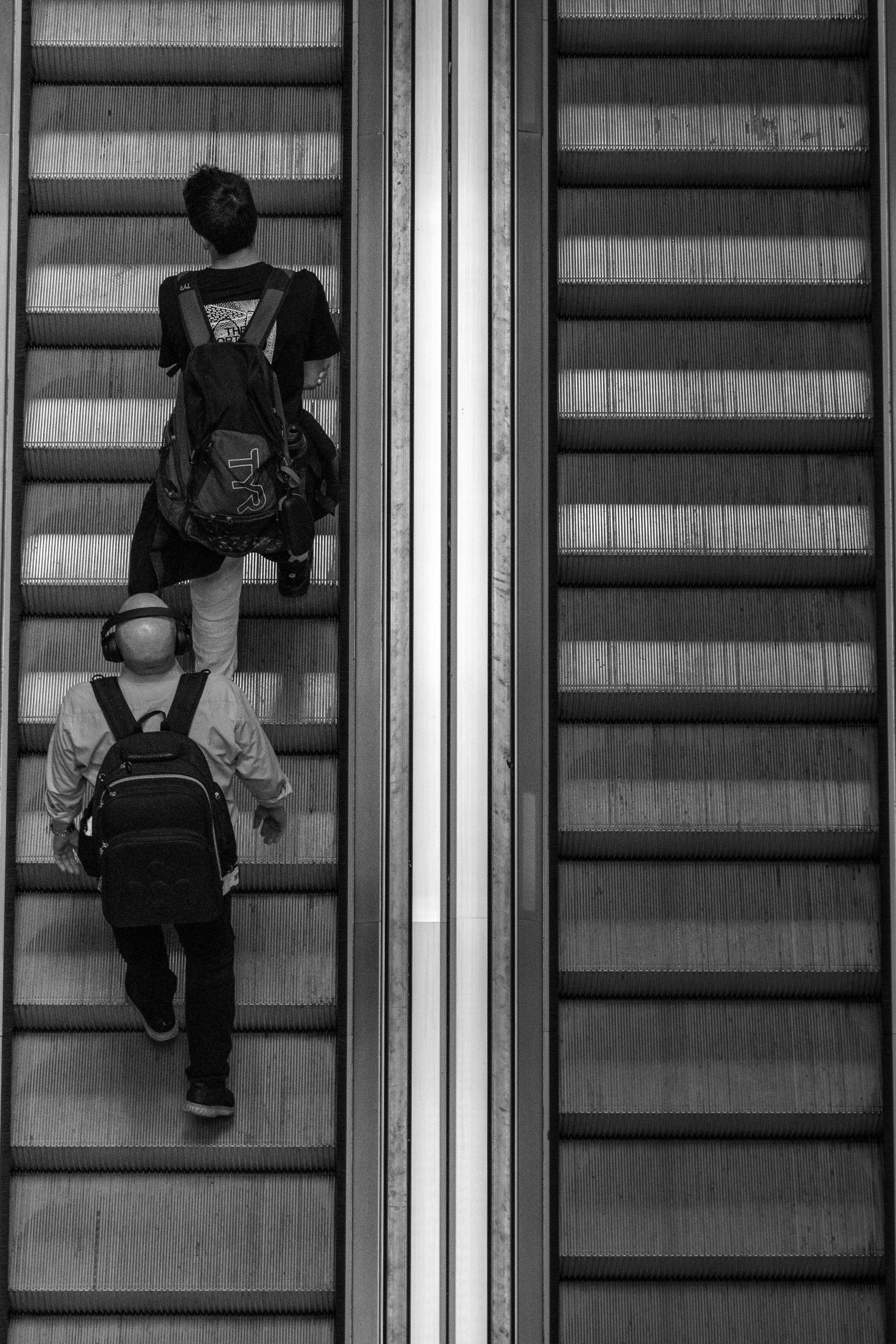 Two men with backpacks ascend an escalator in a monochrome street photo.