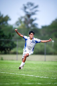 Young soccer player in white and blue kit celebrating a goal on a green field.