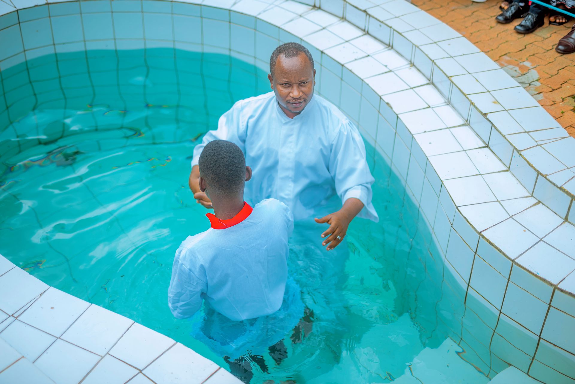 A religious baptism ceremony taking place in a tiled pool with two participants.