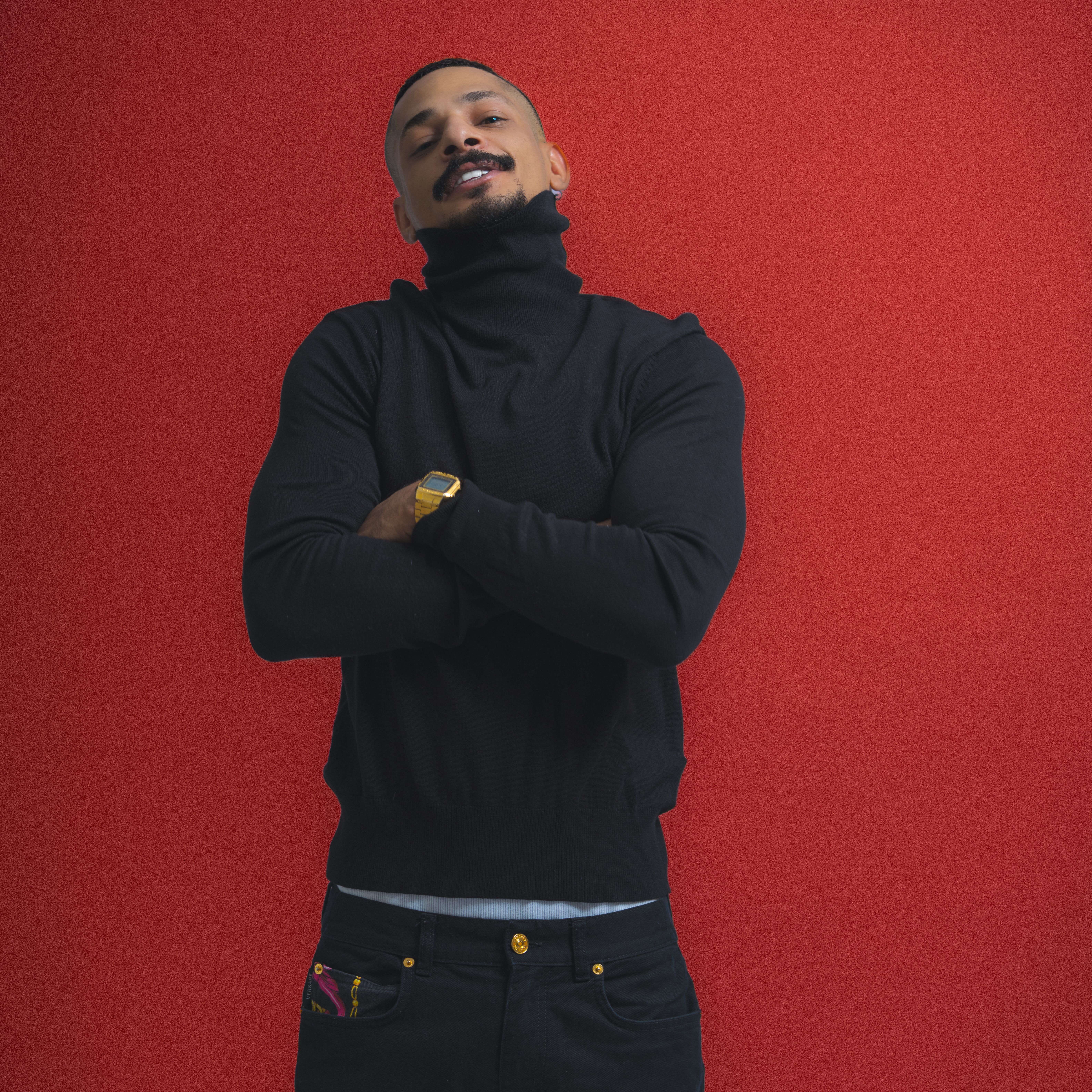 Fashionable man wearing black with watch, confidently posing in front of red background.