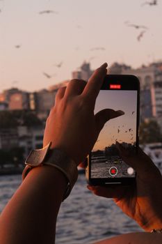 Person records sunset over city skyline with a smartphone, seagulls flying.