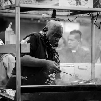 Black and white photo of a street vendor preparing food at an outdoor market with focused expression.