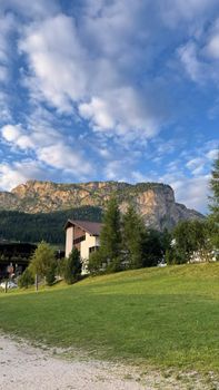 Idyllic rural setting with a house on a green hill, framed by majestic mountains and blue sky.