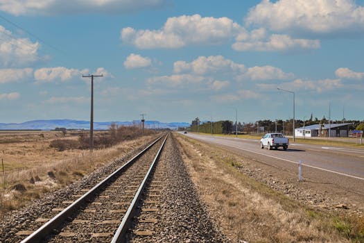 Scenic view of a railway track and rural road under a blue sky with clouds.