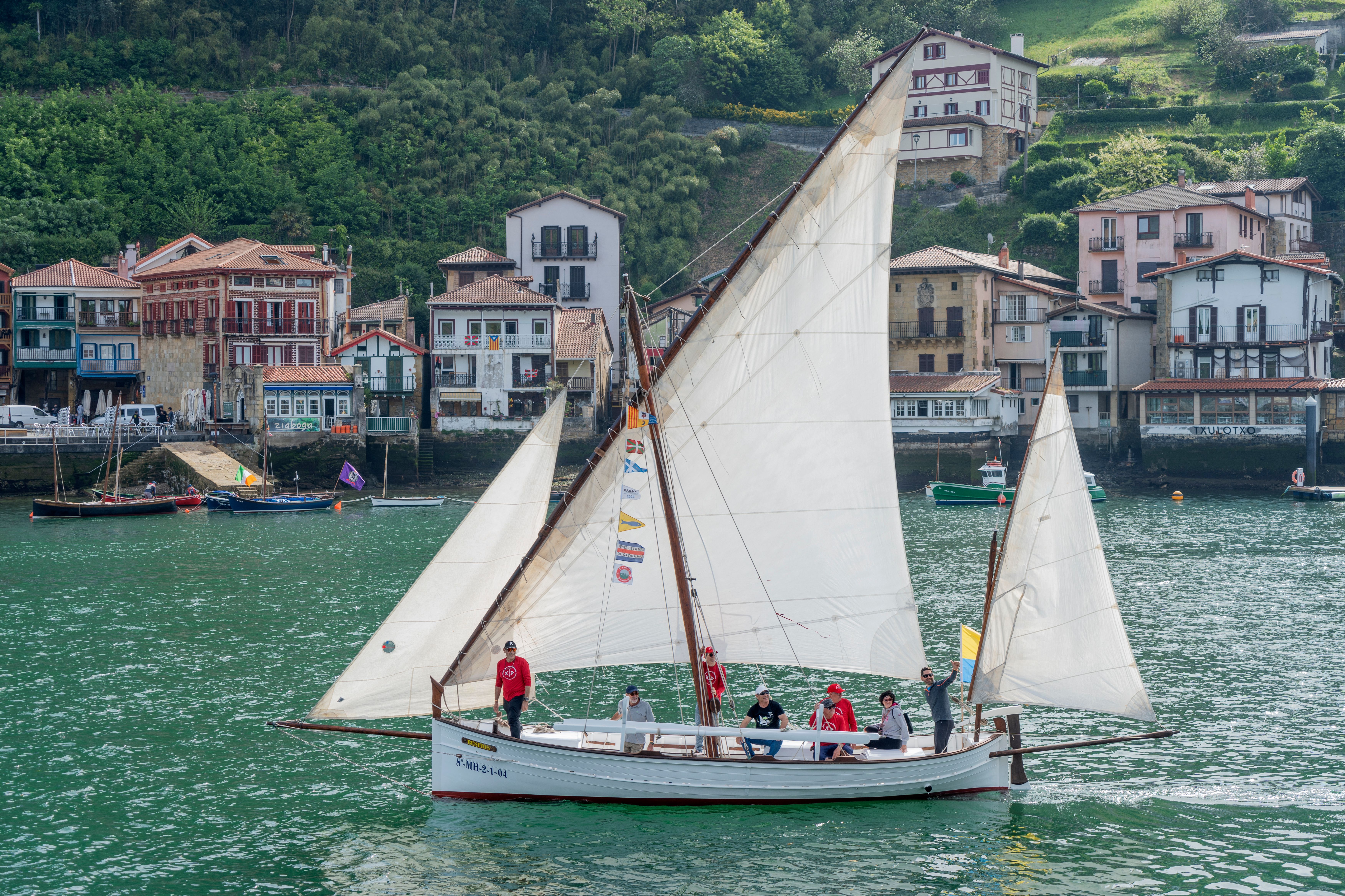 Traditional sailboat with crew sailing near a scenic coastal village, surrounded by lush green hills.