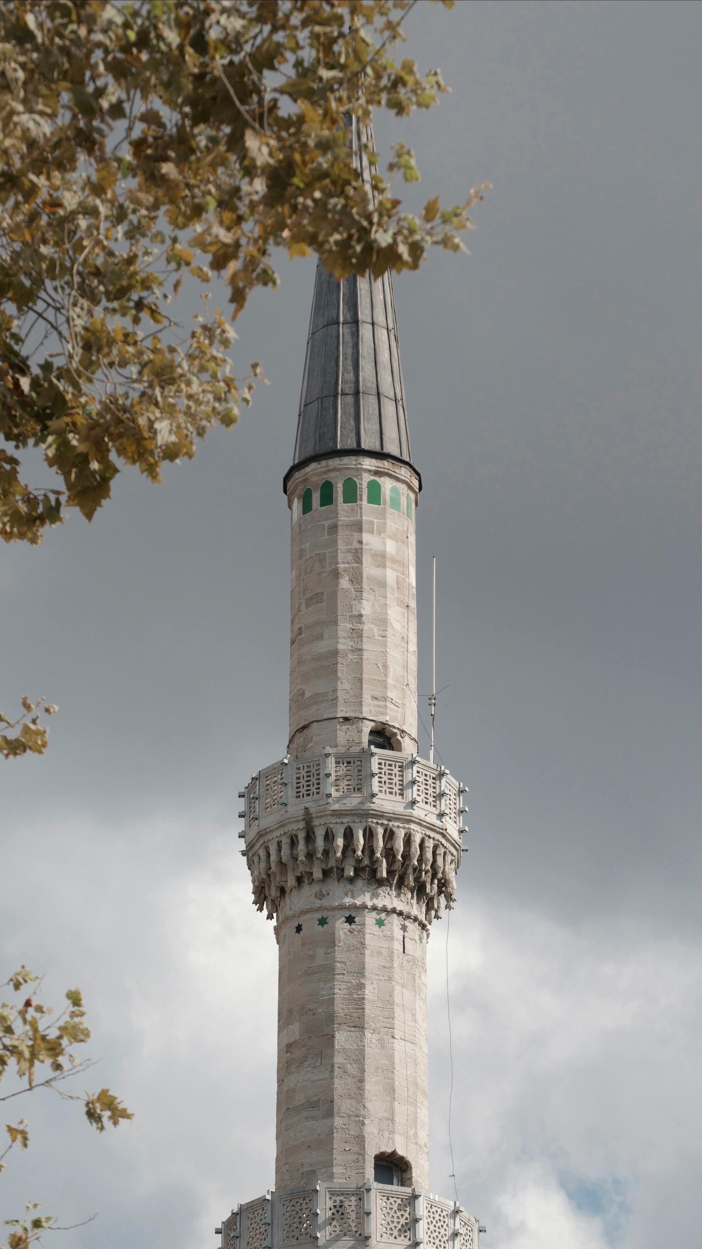 A tall minaret from a mosque in Istanbul with a cloudy backdrop and tree foliage.