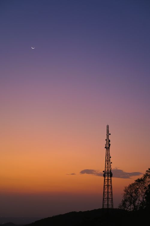 Free Communication tower under a vibrant orange sunset sky with crescent moon. Stock Photo