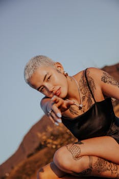 A stylish woman with tattoos and a short hairstyle poses outdoors during sunset, exuding confidence and glamour.