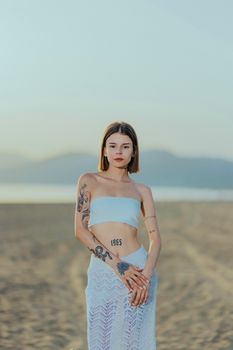 Serene image of a young woman with tattoos standing on a sandy beach at sunset.