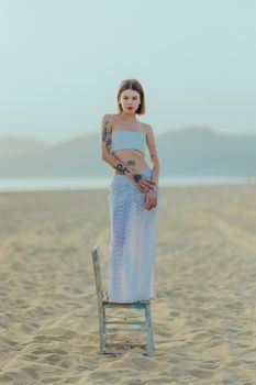 Stylish woman in a summer dress standing on a beach chair, exuding confidence and grace on a tranquil sandy beach.