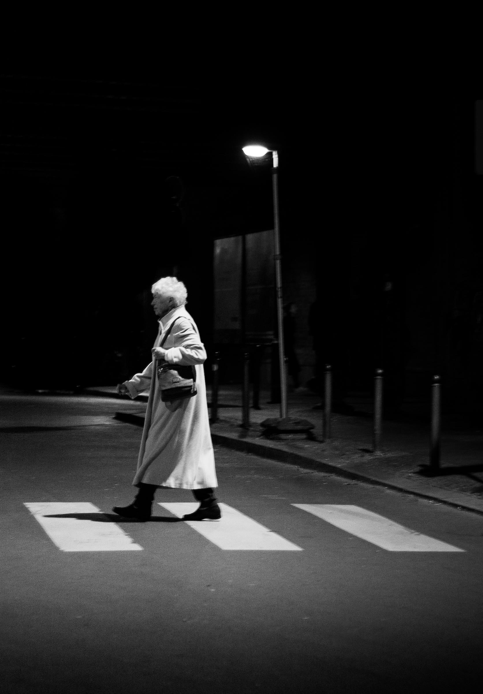 Black and white photo of a senior woman crossing a street at night under a streetlamp.